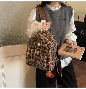 Detail view of leopard print plush backpack with gold logo plate and fuzzy pom-pom charm