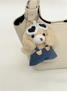 Close-up of a plush beige bear charm with denim overalls hanging on a cream bucket bag