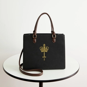Classic black tote bag with gold crown crest and adjustable brown straps on white desk