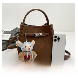 Brown lychee leather bucket bag with bear pendant and adjustable strap next to a smartphone