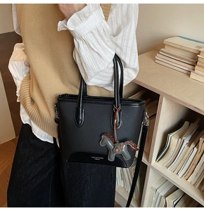 Detalle de un bolso cubo negro con asas y colgante de caballo frente a una estantería
