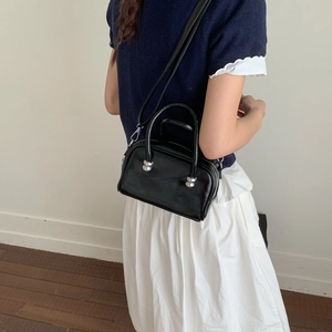 Sac bandoulière en cuir brillant noir porté par un mannequin avec une jupe plissée