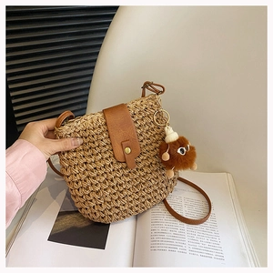 Detail of woven straw shoulder bag featuring brown leather flap and plush ball charm