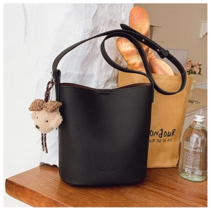Close-up of black litchi texture bucket bag with cute plush bear charm on wooden desk