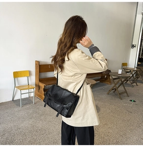 Black leather messenger bag with flap closure and metal rings worn with a beige trench coat