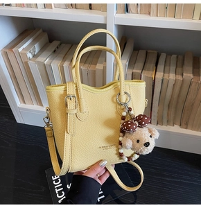 Detail of pale yellow pebbled leather bucket bag with cute plush bear charm on a bookshelf