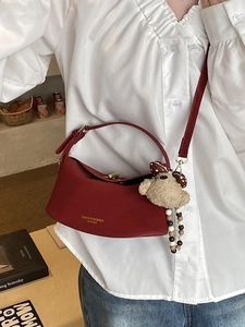 Deep red half-moon shoulder bag with plush bear charm worn by a model in a cozy setting