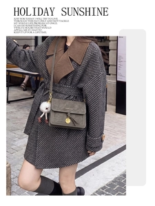 Detail of a dark green crossbody bag with a round buckle and plush pendant on a grey coat