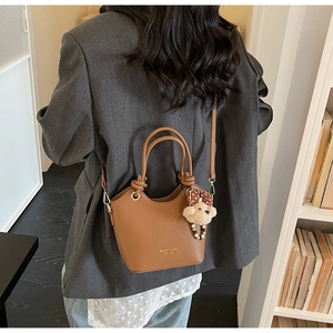 Brown litchi grain bucket bag with dual handles and teddy bear charm on grey blazer
