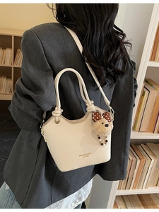 Minimalist beige bucket bag with teddy bear charm and twin handles on a bookshelf background