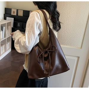 Brown leather tote bag with decorative bow shown on shoulder in cozy library setting
