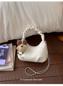 Detail of white half-moon bag featuring pleated handle and teddy bear charm on wood
