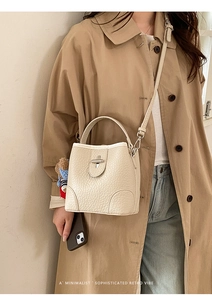 Minimalist beige pebble leather bucket bag with top handle and plush charm on trench coat
