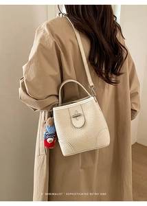 Close-up of a minimalist cream bucket bag featuring textured faux leather and a bear charm