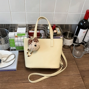 Cream trapezoid handbag with plush bear charm on dark wood table in home setting