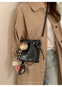 Detail of black quilted bucket bag with gold chain and plush bear charm on camel coat