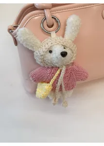 Close-up of a white plush bear charm in pink knit sweater hanging on a shell-shaped handbag