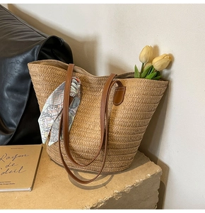 Detail of hand-woven straw tote bag featuring brown leather handles and a green silk scarf