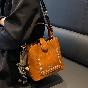 Brown litchi grain bucket bag with wide strap and cute rabbit pendant on a leather bench