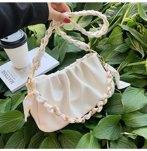 Detail van gebroken witte cloud bag met gevlochten handvat en zijden sjaalketting op groene bladeren