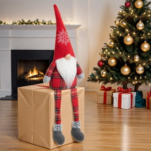 Red Christmas gnome plush with long legs sitting on gift box by a cozy fireplace