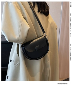 Close-up of black saddle crossbody bag with metal hooks on a trench coat background