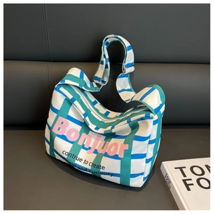 Canvas shoulder bag with green checkered stripes and pink Bonjour text on leather sofa