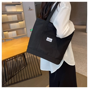 Model wearing a large black canvas tote bag with side reinforcement straps and brand tag