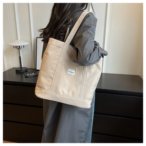 Detail of beige canvas tote bag with reinforced straps and logo patch worn by a model