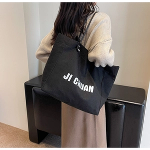 Large black canvas tote bag with white lettering worn by a model in a natural setting