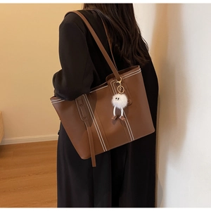 Detail of a brown leather tote bag with white stitching and a plush doll pendant
