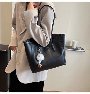 Large black leather tote bag with plush charm shown in a minimalist commuter setting