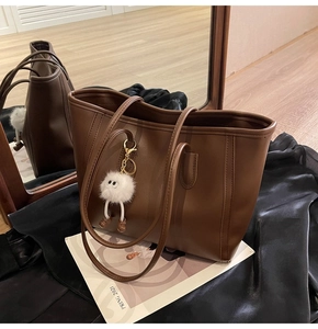 Detail of a large brown faux leather tote bag featuring a white plush pompom charm decoration