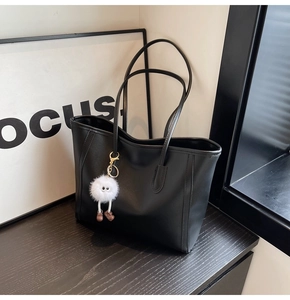 Detail of a large black leather tote bag with a white fluffy monster charm on a wood table