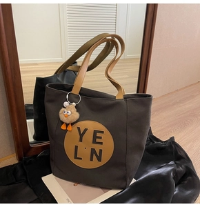 Close-up of a charcoal grey canvas tote bag featuring tan leather handles and a plush bird charm
