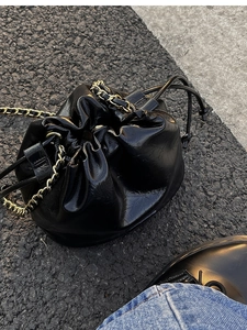 Detail of black glossy drawstring bucket bag with gold-tone chain strap on asphalt