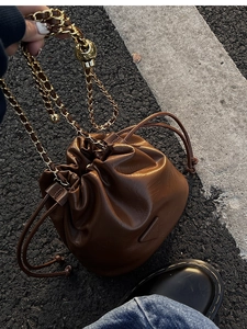 Detail of dark brown bucket bag with gold chain strap and drawstring closure on asphalt