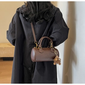 Detail of dark brown cylinder Boston bag with embossed logo and fuzzy ball charm on black coat
