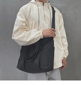 Model wearing a large black nylon tote bag with wide strap on a grey wall background