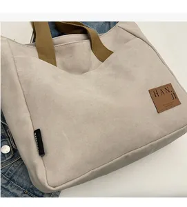 Detail of canvas tote bag with brown woven handles and leather logo patch on white surface