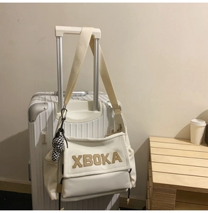 Close-up of cream tote bag with XBOKA lettering and checkered charm on a suitcase