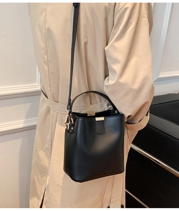 Model wearing a black bucket bag with a beige coat against a minimalist wall background