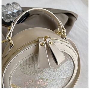 Detailed close-up of beige round handbag with floral embroidery and gold zipper detail