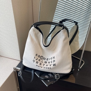 Minimalist white canvas tote bag with black alphabet design and leather strap shown in sunlight