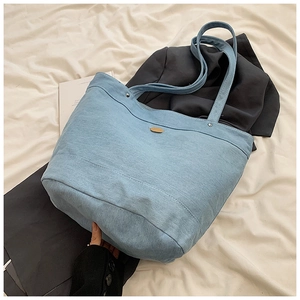 Detail of light blue denim tote bag with metallic oval logo on soft fabric surface