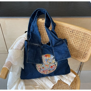 Détail de la broderie sur un grand sac en jean bleu posé sur une chaise en osier rétro