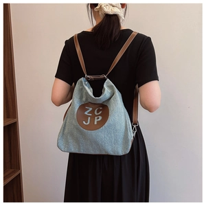 Detail of light blue denim backpack with brown leather strap and circular letter patch