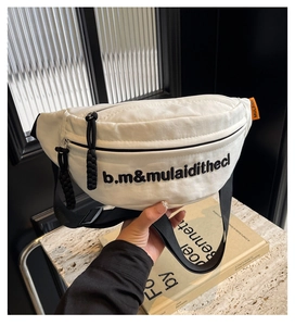 Detailaufnahme der weißen Canvas-Brusttasche mit schwarzer Stickerei und orangem Etikett