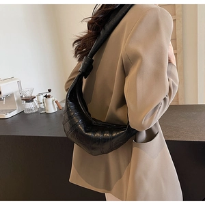 Model wearing a black glossy pleated croissant bag over a camel blazer in a minimalist cafe