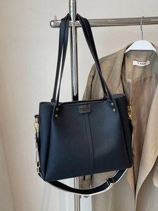 Large black leather tote bag with wide strap and gold rivets on a khaki trench coat background